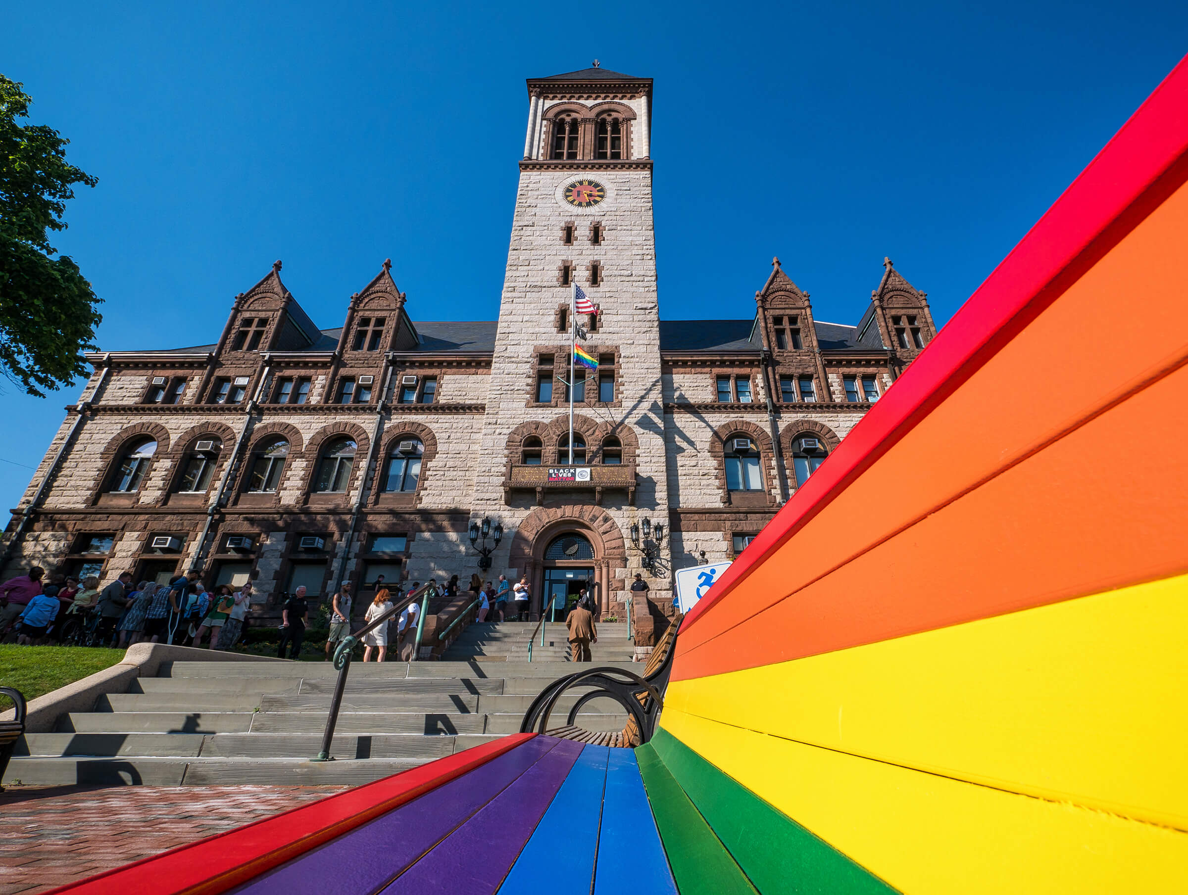 A historic stone city hall building in Cambridge MA with a clock tower stands under a clear blue sky. In the foreground, a rainbow-colored bench symbolizes LGBTQ+ Pride.