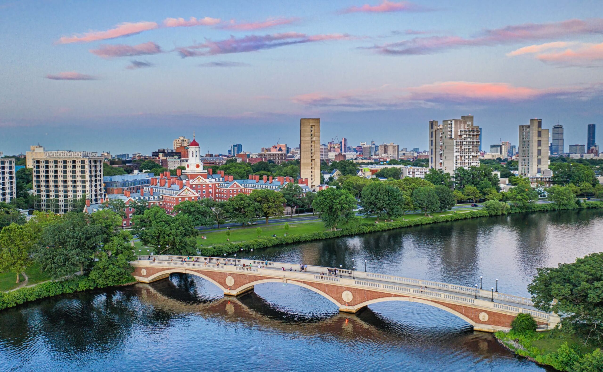 A scenic aerial view of the Charles River in Cambridge, Massachusetts, with the historic John W. Weeks Footbridge crossing the water. In the background, the iconic red-domed Harvard University building and a mix of modern and historic architecture can be seen. The sky is painted with soft pastel clouds during sunset, reflecting beautifully on the river below.
