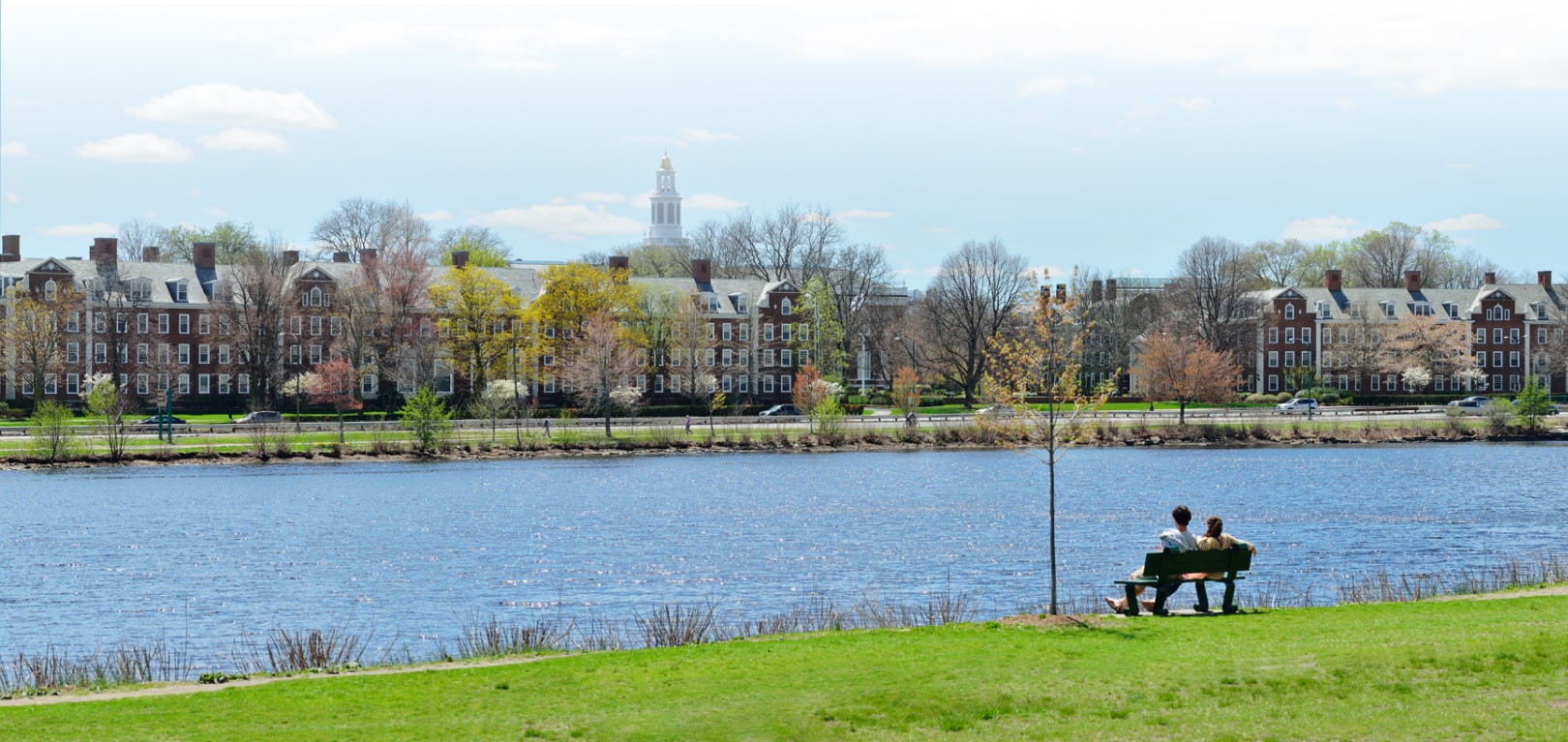 Wide landscape view of the Charles River. A couple sits on a park bench, taking in the tree-lined Cambridge cityscape on the opposite bank.