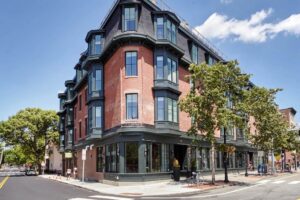 A modernized historic brick building with large bay windows and black trim sits on a sunny street corner, surrounded by trees, bicycles, and a few pedestrians.