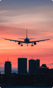 A plane glides into Boston airport, silhouetted against a pink and blue sunset with the Boston, MA skyline in the background.