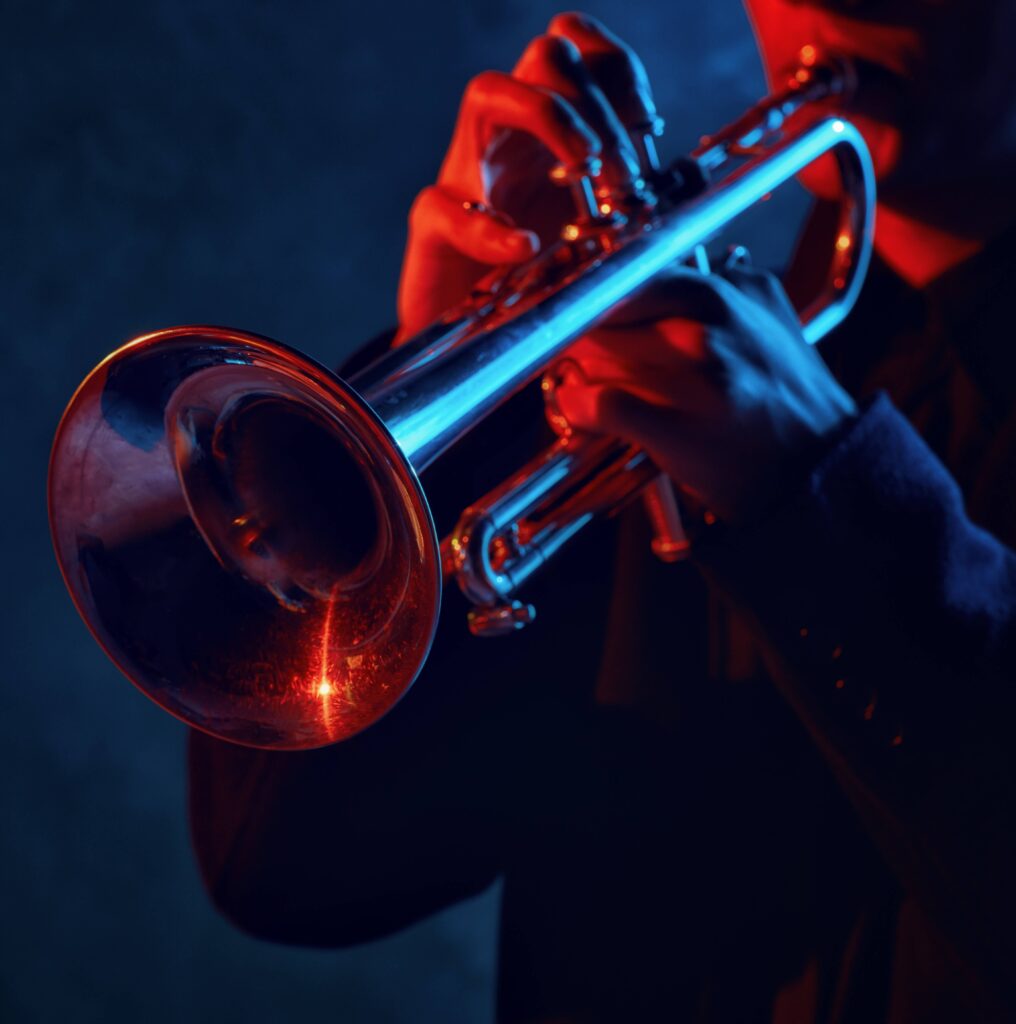 Close-up of a musician playing a trumpet, illuminated by dramatic red and blue stage lighting, highlighting the shiny metal surface of the instrument and the player’s focused grip.