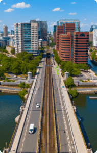 Aerial view of a bridge highway leading to the city center of Cambridge, MA. Tall buildings stand out against a bright blue sky.