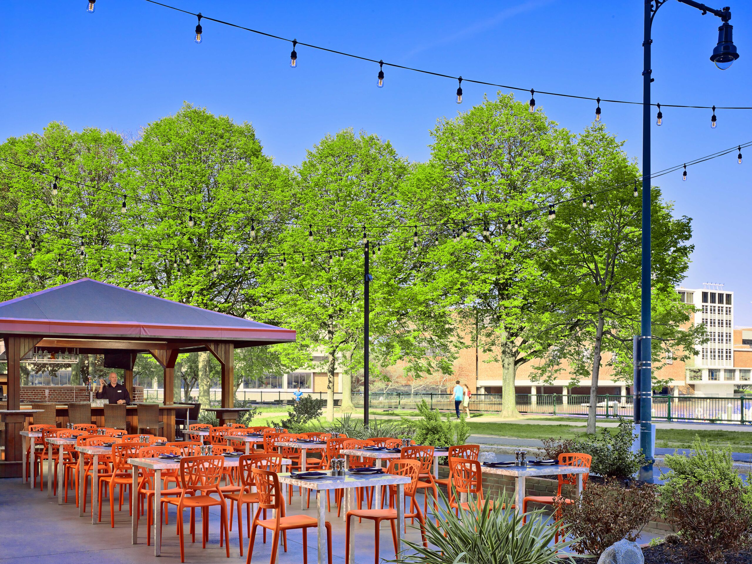 A bright outdoor patio dining area featuring modern orange chairs and white tables arranged neatly under string lights. A wooden bar pavilion stands to the left, surrounded by lush green trees and a clear blue sky, creating a relaxed, inviting atmosphere.