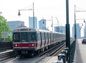 A red and silver subway train travels across a bridge into the city with modern skyscrapers in the background, passing street lamps and running parallel to a road with a red car and a few pedestrians