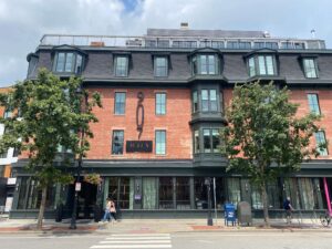 A brick building with dark green trim and large bay windows, located at 907 Main Street. The lower level has large glass windows and an entrance with a sign reading "Blue Owl." Two trees line the sidewalk in front, and pedestrians are walking by. A USPS mailbox and newspaper stand are also visible near the street.